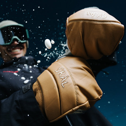 A person in winter gear wearing a tan SKAL ski glove in an outdoor setting, with opening a canned drink.