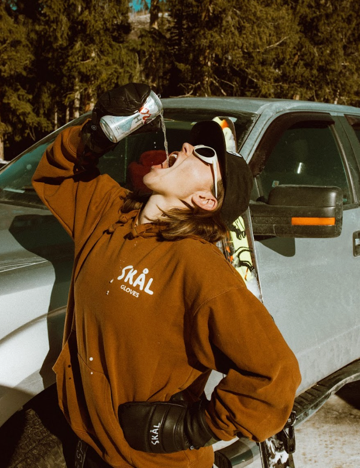 Person in a brown jacket with 'SKØL' logo drinking from a metal cup outdoors.